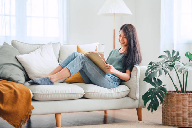 Young asian woman sitting on modern sofa in front of window relaxing in her living room reading book and drinking coffee or tea at home Young asian woman sitting on modern sofa in front of window relaxing in her living room reading book and drinking coffee or tea at home Reading stock pictures, royalty-free photos & images